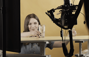 website2027-Bilder Kopie3 - fokusmedien Woman smiling at a glass of water on a studio table, with a large video camera in the foreground.