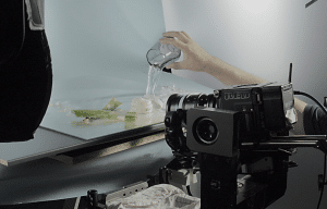website2027-Bilder Kopie7 - fokusmedien Behind-the-scenes shot of a RED cinema camera on a rig as a hand pours water into a glass on a light blue backdrop for a splash shot.