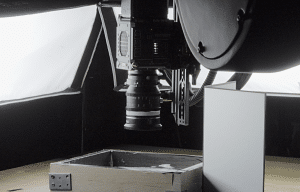 website2027-Bilder Kopie9 - fokusmedien Close-up of a camera/lens mounted on a rig over a wooden table with a metal tray, in a black photography setup tent.