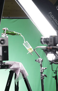 website2027-BilderV - fokusmedien Beer bottle pouring into a glass held by a clamp on a stand during a studio shoot, with bright lights and a green screen backdrop.